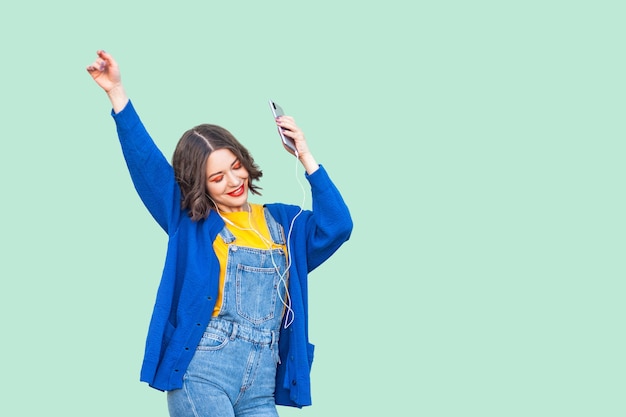 Bella giovane ragazza adulta positiva in hipster indossare in tuta di jeans in piedi, tenendo il telefono e ascoltando la musica preferita con i telefoni e ballando. Studio girato, sfondo verde, isolato