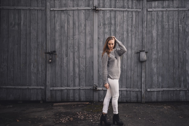 Bella giovane ragazza bionda di modello. Pantaloni bianchi. maglione lavorato a maglia grigio. stivali neri. ciondolo in legno sul collo a forma di cavallo.