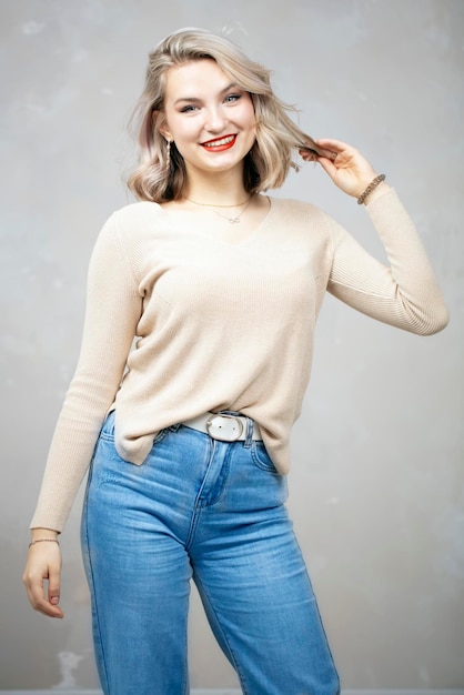 Bella giovane ragazza bionda in jeans e un maglione beige che posa alla macchina fotografica