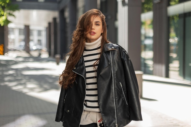 Bella giovane ragazza bruna alla moda in abiti alla moda con un'elegante giacca di pelle e un maglione a righe cammina in città in una giornata di sole