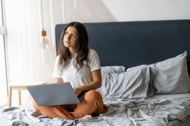 Bella giovane ragazza castana con un computer portatile che si siede sul letto