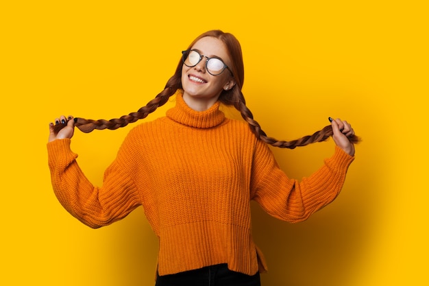 Bella giovane ragazza caucasica con i capelli rossi che tiene le sue code vestita con un maglione arancione in posa su uno sfondo giallo