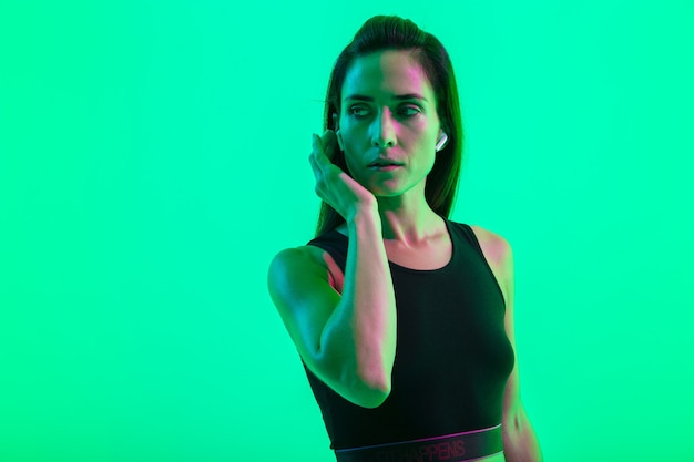 Bella giovane ragazza fitness in piedi isolata su una parete al neon verde, con indosso auricolari wireless