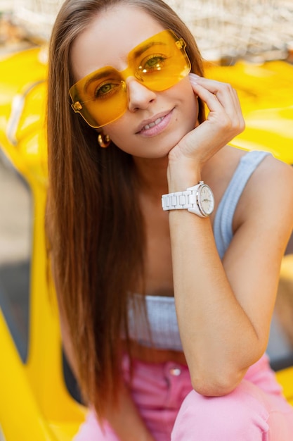 Bella giovane ragazza hippie felice in abiti luminosi alla moda con occhiali da sole gialli alla moda alla moda in top blu e denim rosa seduto su un'auto gialla colorata vintage e guarda alla telecamera