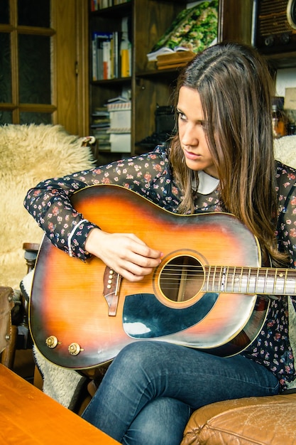 Bella giovane ragazza hipster che suona la chitarra acustica seduta in un divano a casa. Edizione a colori vintage retrò