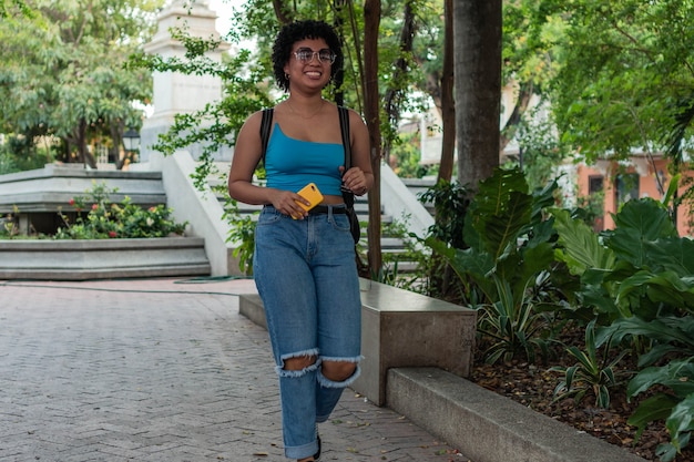 Bella giovane ragazza ispanica che cammina all'aperto nel parco, portando lo zaino, usando il cellulare