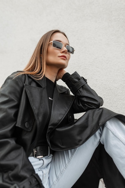 Bella giovane ragazza modello caucasica con occhiali da sole vintage oro in un cappotto di pelle nera alla moda e jeans seduti sulla strada