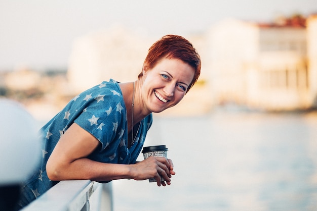 Bella giovane ragazza sorridente dai capelli rossi con i capelli corti sul molo in camicetta di jeans e gonna ritratto all'aperto.