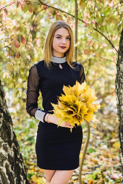 Bella giovane studentessa in abito nero che tiene un mazzo di foglie nel parco autunnale.