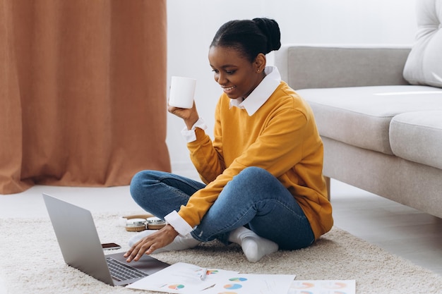 Bella giovane studentessa universitaria nera seduta sul pavimento, bevendo caffè e facendo progetti sul computer portatile