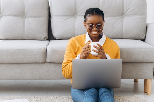 Bella giovane studentessa universitaria nera seduta sul pavimento, bevendo caffè e facendo progetti sul computer portatile