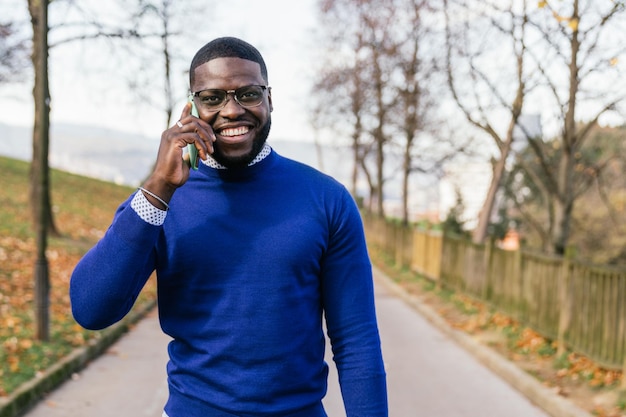Bella gioventù africana in maglione blu casual che irradia sorriso e occhiali che chiacchierano al telefono in un parco ben illuminato
