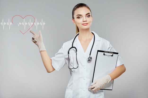 Bella infermiera che indossa veste medica bianca e stetoscopi a sfondo grigio, assistenza sanitaria e concetto di farmacologia, ragazza in uniforme bianca, indicando con la mano al cardiogramma