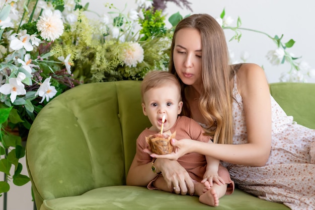 Bella madre amorevole in un vestito siede insieme al figlio piccolo su un divano verde circondato da bellissimi fiori Tiene un cupcake con una candela in mano Il ragazzo urla con la bocca spalancata