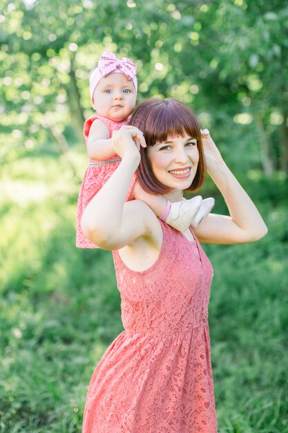 Bella madre con il cappello di paglia E la sua piccola figlia all'aperto la famiglia cerca in un vestito rosa. aspetto familiare. bambino piccolo, una bambina si siede sulle spalle della mamma sorridendo