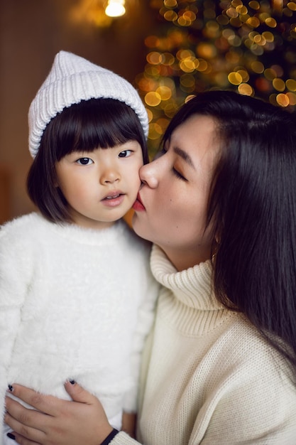 Bella mamma coreana e figlia del bambino che si siedono in maglioni a maglia bianchi all'albero di Natale nello studio d'oro