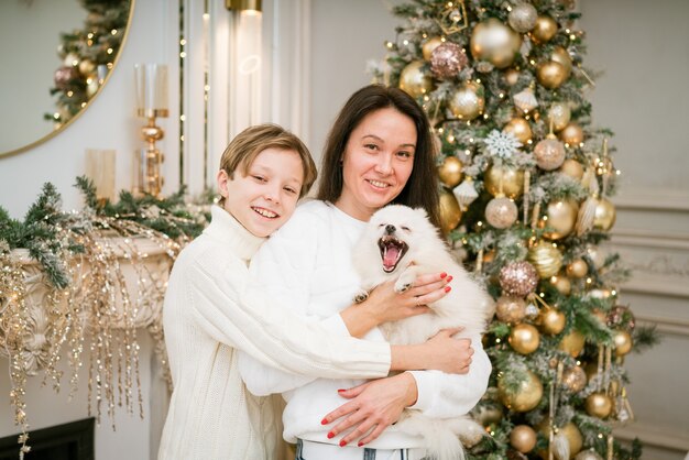 Bella mamma sorridente e figlio che si abbracciano con un cane bianco felice a casa per le vacanze di natale felice fa...