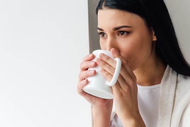 Bella mattina. Ritratto di bella giovane donna che beve caffè e guarda attraverso la finestra mentre si trova vicino al muro bianco white