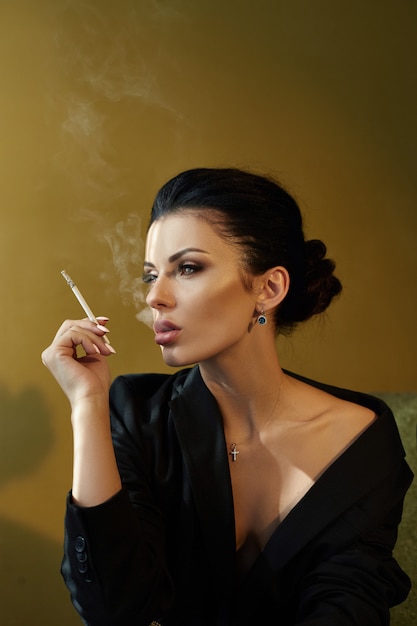 Bella moda Donna nuda fiduciosa con i capelli neri Fumare una sigaretta seduto su una sedia in una giacca nera