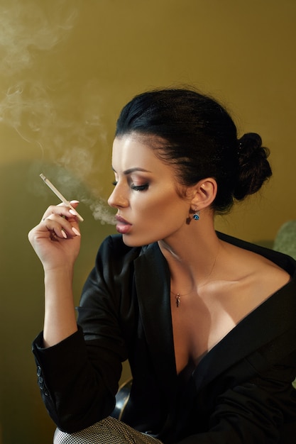 Bella moda Donna sicura nuda con i capelli neri Fumare una sigaretta seduto su una sedia