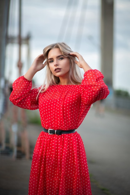 Bella modella bionda che indossa un abito rosso alla moda, in posa sul ponte la sera