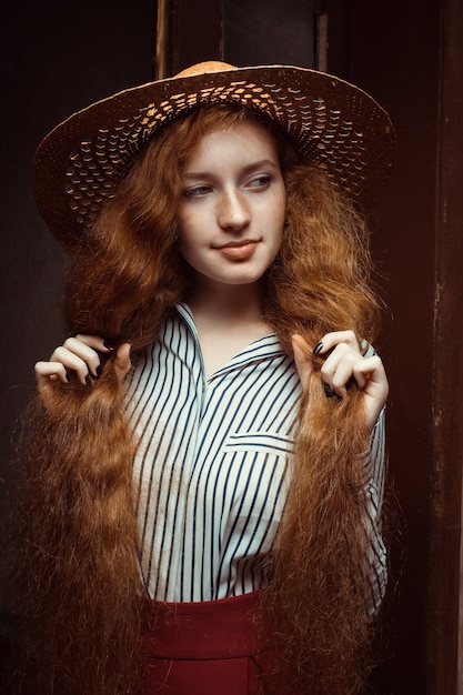 Bella modella rossa con lunghi capelli ricci in cappello di paglia in posa vicino alla vecchia porta di legno