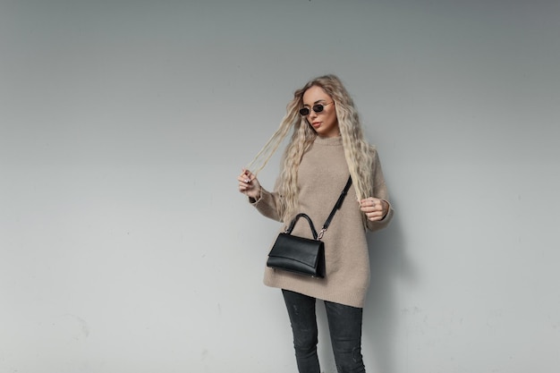 Bella modella urbana alla moda con un'acconciatura bionda riccia con occhiali da sole vintage in un elegante maglione beige con una borsa nera alla moda si trova vicino a un muro grigio in città