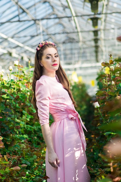 Bella modella vestita con un lungo abito rosa posa tra i tanti fiori della serra