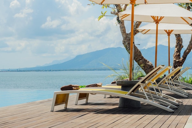 Bella piscina tropicale in hotel o resort con ombrellone, noci di cocco, albero, lettini, palme, con vista sulla piscina a sfioro, sull'oceano e sullo sfondo delle montagne