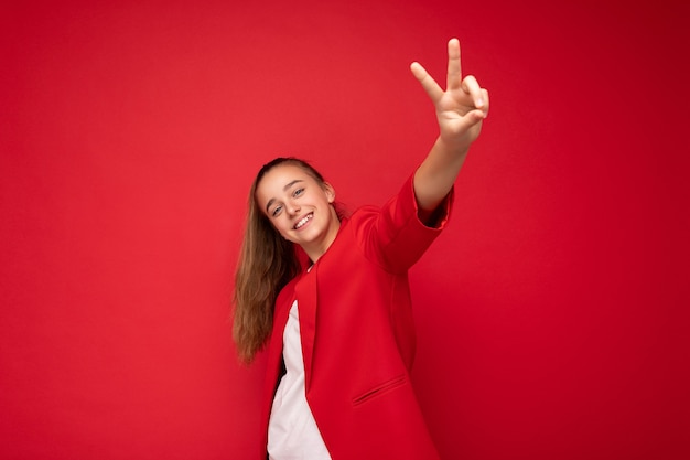 Bella positivo felice sorridente bruna bambina indossa giacca rossa alla moda e t-shirt bianca in piedi isolato su sfondo rosso muro guardando la fotocamera e mostrando gesto di pace