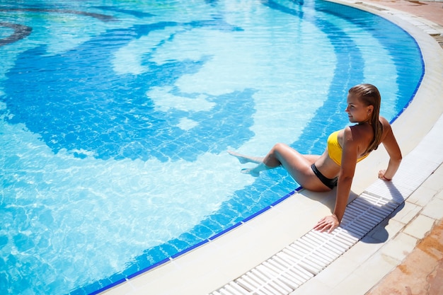 Bella ragazza abbronzata in costume da bagno giallo in una grande piscina con acqua blu