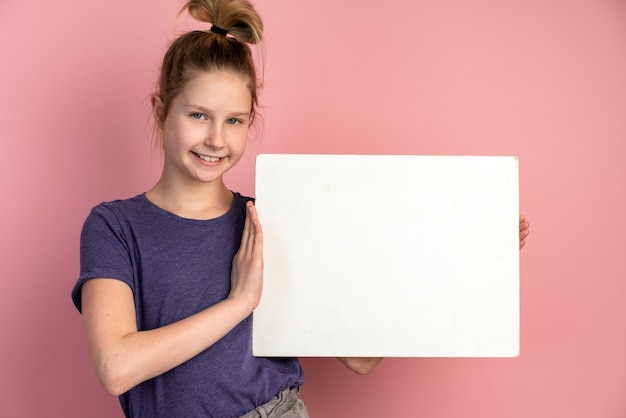 Bella ragazza adolescente bionda con gli occhi azzurri tiene un foglio di carta bianco su una parete rosa e sorride