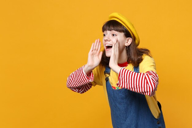 Bella ragazza adolescente in berretto francese che guarda da parte, sussurra pettegolezzi e racconta il segreto con il gesto della mano isolato su sfondo giallo. Persone sincere emozioni, concetto di stile di vita. Mock up spazio di copia.