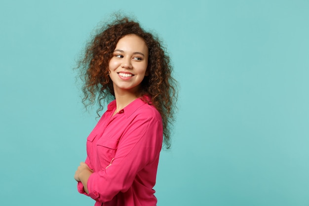 Bella ragazza africana in abiti casual rosa che si tiene per mano piegata, guardando da parte isolata sul fondo della parete turchese blu in studio. Persone sincere emozioni, concetto di stile di vita. Mock up copia spazio.