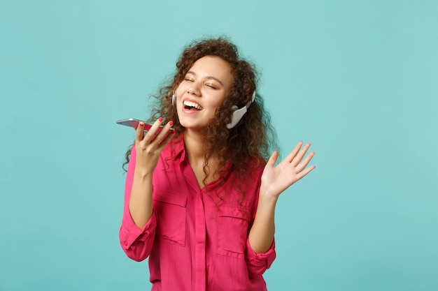 Bella ragazza africana in abiti casual tiene il telefono cellulare, ascoltando musica con le cuffie isolate su sfondo blu turchese in studio. Concetto di stile di vita di persone sincere emozioni. Mock up copia spazio.