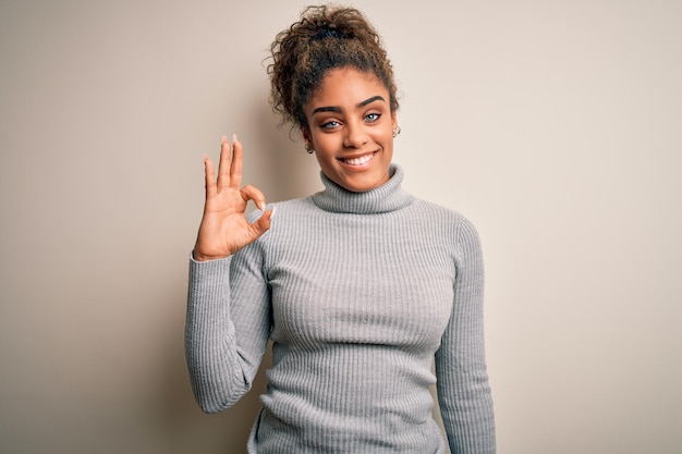 Bella ragazza afroamericana che indossa un maglione a collo alto in piedi su sfondo bianco sorridente positivo facendo segno ok con la mano e le dita Espressione di successo