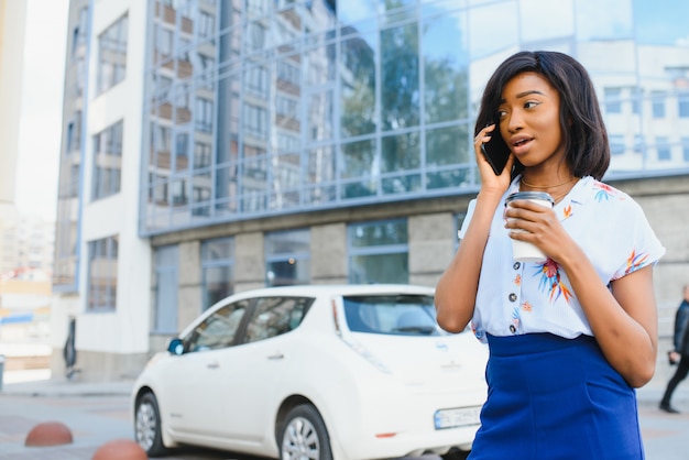 Bella ragazza afroamericana che sta sulla via con il cellulare e caffè in mani mentre felicemente guardando da parte.