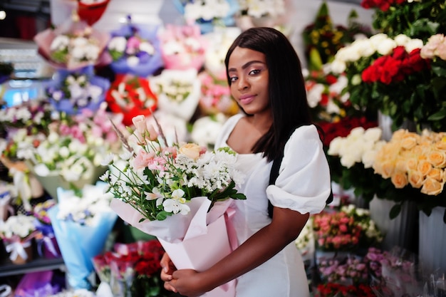 Bella ragazza afroamericana in tenero abito bianco con bouquet di fiori in mano in piedi su sfondo floreale nel negozio di fioriFioraio nero