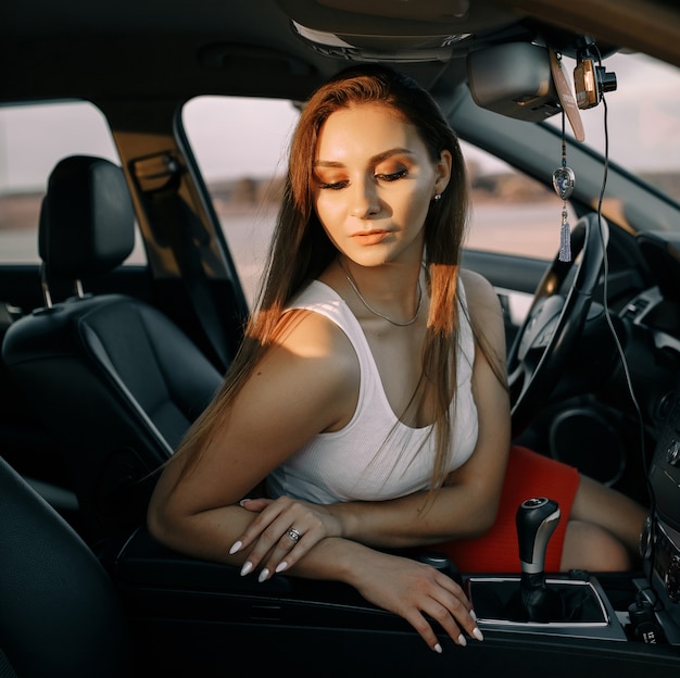 Bella ragazza alla guida di un'auto la sera sotto il sole al tramonto su un parcheggio vuoto