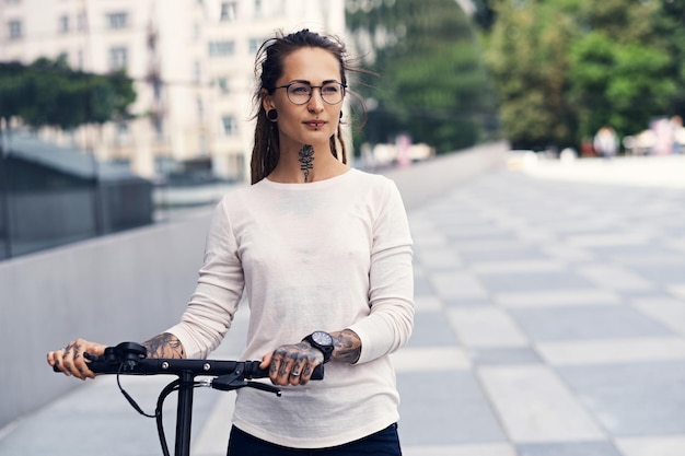 Bella ragazza alla moda con i dreadlocks sta guidando uno scooter elettrico per strada in una nuvolosa giornata estiva.