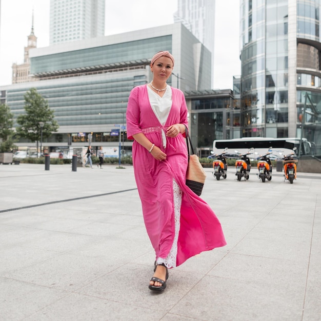 Bella ragazza alla moda con una fascia in abiti estivi luminosi alla moda con un pareo rosa e pantaloni di pizzo bianco con una borsa di paglia intrecciata che cammina nella città moderna