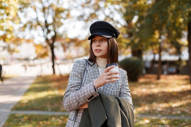 Bella ragazza alla moda in un cappello e vestito a scacchi di moda con caffè passeggiate nel parco in una giornata di sole autunnale