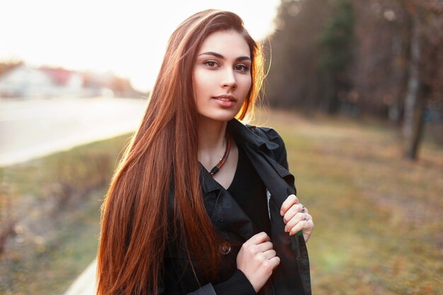 Bella ragazza alla moda in un mantello nero che cammina nel parco in una giornata di primavera al tramonto