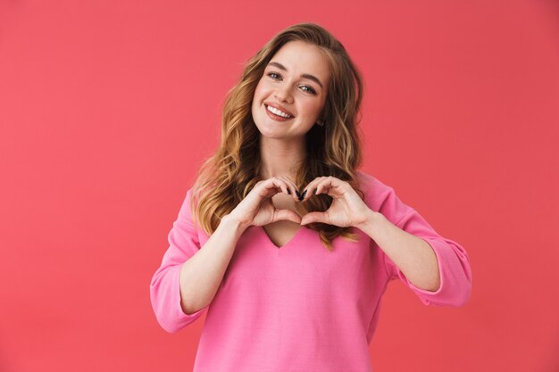 Bella ragazza allegra che indossa abiti casual in piedi isolata sul muro rosa, mostrando il gesto del cuore