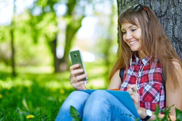 Bella ragazza allegra con uno smartphone seduto in un parco su un prato in una giornata di sole adolescente