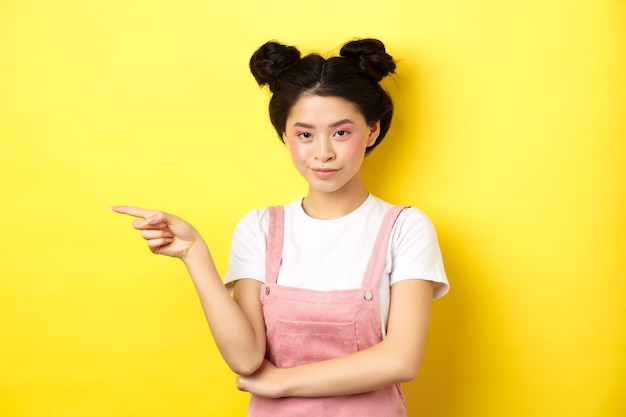 Bella ragazza asiatica adolescente con trucco luminoso, dito puntato a sinistra al banner e sorridente, giallo.