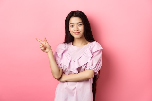 Bella ragazza asiatica adolescente puntare il dito a sinistra, sorridente fiducioso, mostrando pubblicità su sfondo rosa.