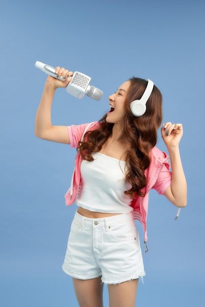 Bella ragazza asiatica con microfono canta e cuffie isolate su sfondo blu.