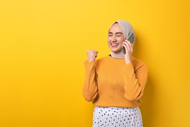 Bella ragazza asiatica eccitata che indossa l'hijab parlando al cellulare ottenendo buone notizie alzando il pugno dicendo sì isolato su sfondo giallo