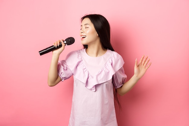 Bella ragazza asiatica esegue la canzone, cantando nel microfono e sorridente romantico, in piedi in abito su sfondo rosa.